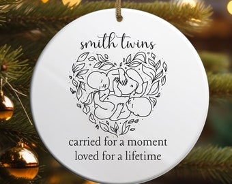 May include: White ceramic ornament with a black and white illustration of two babies in a heart shape with leaves. The text "smith twins" is at the top and "carried for a moment loved for a lifetime" is at the bottom.