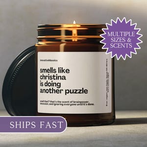 Puede incluir: Un tarro de vela de vidrio marrón encendido con una tapa negra. La etiqueta dice "smells like christina is doing another puzzle". Una pegatina morada dice "Multiple Sizes & Scents".
