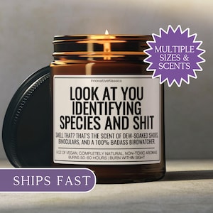 May include: A brown glass jar candle with a black lid, featuring a white label with black text that reads "LOOK AT YOU IDENTIFYING SPECIES AND SHIT." The candle has a lit wick and a purple sticker that says "MULTIPLE SIZES & SCENTS."