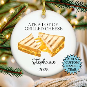 May include: White ceramic Christmas ornament with a gold string. The ornament features a watercolor illustration of two grilled cheese sandwiches and the text "ATE A LOT OF GRILLED CHEESE". Includes a name and the year 2025.
