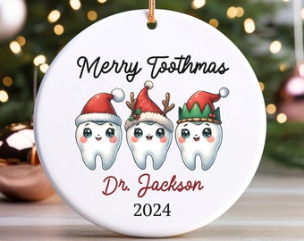 May include: White ceramic Christmas ornament with three cartoon teeth wearing Santa hats, reindeer antlers, and an elf hat. The ornament says "Merry Toothmas" and "Dr. Jackson 2024".