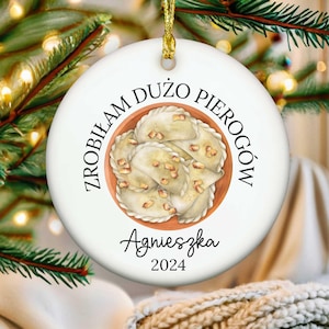 May include: A white ceramic Christmas ornament with a gold string. The ornament features a watercolor illustration of pierogi in a bowl, with the text "Zrobiłam dużo pierogów" and the name "Agnieszka" with the year "2024".