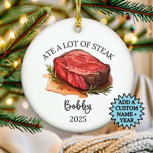 May include: White ceramic Christmas ornament with a gold string. The ornament features a watercolor illustration of a steak with the text "ATE A LOT OF STEAK" above it. Below the steak is the name "Bobby" and the year "2025".