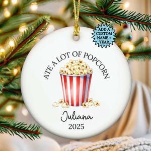 Puede incluir: Adorno navideño redondo de cerámica blanca con una cuerda dorada. El adorno presenta un diseño de cubo de palomitas de maíz a rayas rojas y blancas, el texto "ATE A LOT OF POPCORN", el nombre "Juliana" y el año "2025".