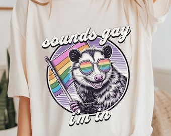 May include: A white t-shirt with a graphic of a possum wearing rainbow sunglasses and holding a rainbow flag. The text "sounds gay i'm in" is printed below the possum.