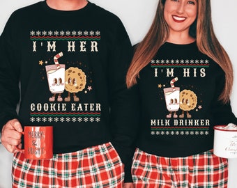 May include: A matching pair of black Christmas jumpers with a milk carton and cookie design. The jumper on the left says "I'm Her Cookie Eater" and the jumper on the right says "I'm His Milk Drinker".