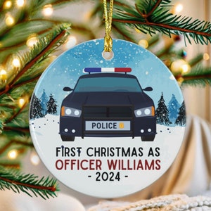 May include: A round, white Christmas ornament with a blue and white winter scene. A black police car with a red and blue light bar is in the foreground. The text "First Christmas as Officer Williams - 2024 -" is printed on the ornament.