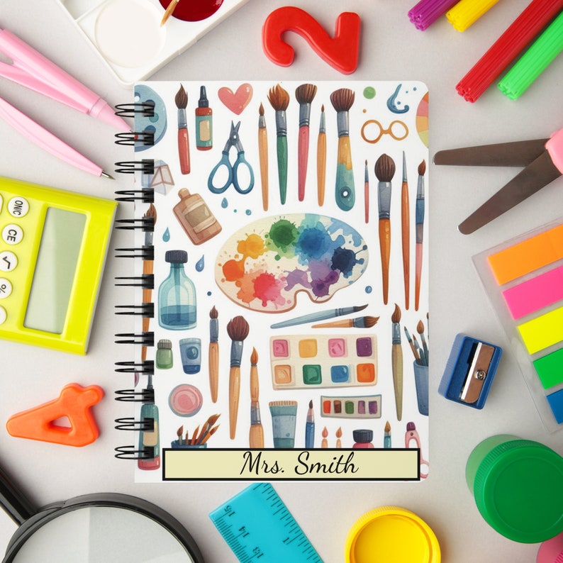 Puede incluir: Una libreta blanca de espiral con un patr&oacute;n colorido de art&iacute;culos de arte. La portada presenta pinceles, paletas de pintura y otras herramientas de arte. La libreta tiene una etiqueta de nombre personalizada que dice "Sra. Smith".