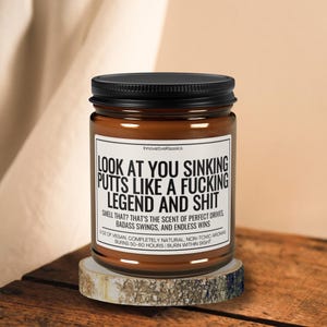 May include: A brown candle in a glass jar with a black lid. The label reads "LOOK AT YOU SINKING PUTTS LIKE A FUCKING LEGEND AND SHIT." The candle sits on a small wooden coaster. The text also mentions the scent and burn time.