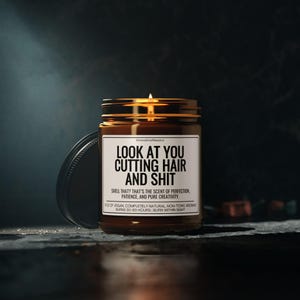 Puede incluir: Una vela encendida en un tarro de vidrio marr&oacute;n con tapa dorada y etiqueta blanca. La etiqueta dice "LOOK AT YOU CUTTING HAIR AND SHIT". La vela est&aacute; sobre una superficie oscura.