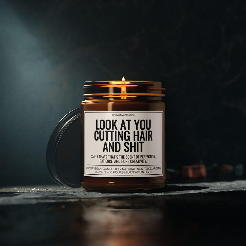 Puede incluir: Una vela encendida en un tarro de vidrio marr&oacute;n con tapa dorada y etiqueta blanca. La etiqueta dice "LOOK AT YOU CUTTING HAIR AND SHIT". La vela est&aacute; sobre una superficie oscura.