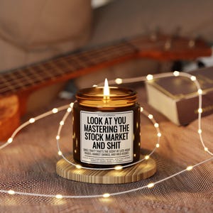 May include: A lit candle in a brown glass jar with a gold lid, sitting on a wooden coaster. The label reads "LOOK AT YOU MASTERING THE STOCK MARKET AND SHIT". Fairy lights and a ukulele are in the background.