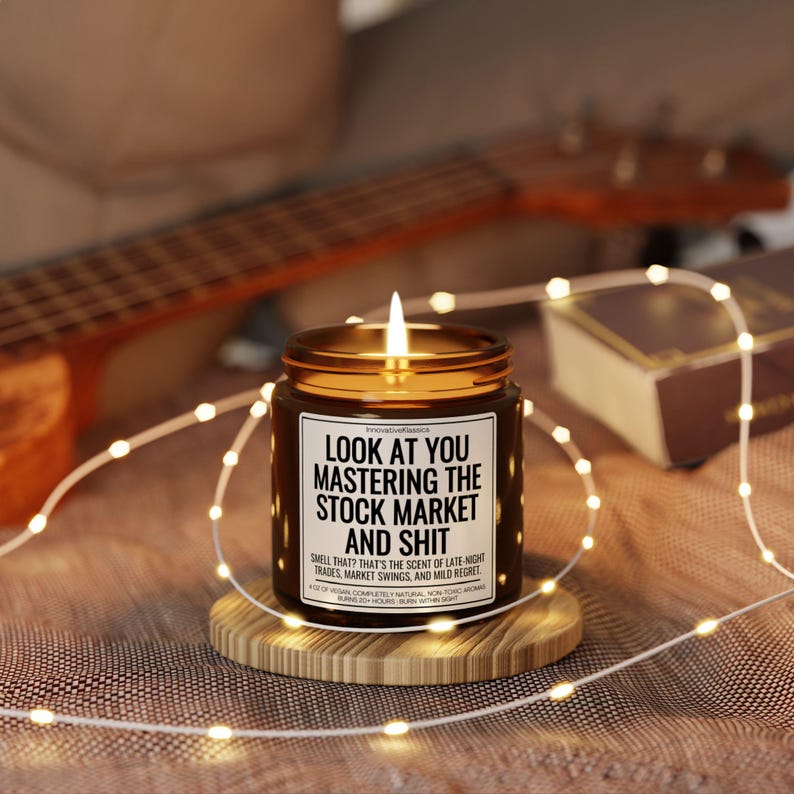 May include: A lit candle in a brown glass jar with a gold lid, sitting on a wooden coaster. The label reads "LOOK AT YOU MASTERING THE STOCK MARKET AND SHIT". Fairy lights and a ukulele are in the background.