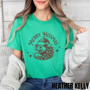 K&ouml;nnte beinhalten: Ein gr&uuml;nes T-Shirt mit einer braunen Illustration eines Opossums, das einen Weihnachtsmann-Hut tr&auml;gt und eine Lichterkette h&auml;lt. Der Text "Merry Hissmas" ist in einem braunen Kreis um das Opossum geschrieben.