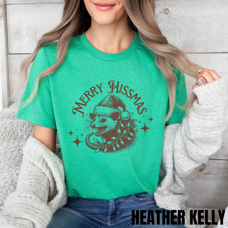 K&ouml;nnte beinhalten: Ein gr&uuml;nes T-Shirt mit einer braunen Illustration eines Opossums, das einen Weihnachtsmann-Hut tr&auml;gt und eine Lichterkette h&auml;lt. Der Text "Merry Hissmas" ist in einem braunen Kreis um das Opossum geschrieben.