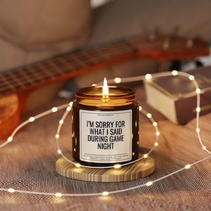 May include: A brown glass candle with a white label that reads "I'm sorry for what I said during game night". The candle is lit and surrounded by fairy lights.