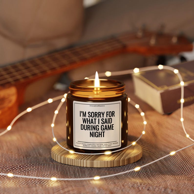 May include: A brown glass candle with a white label that reads "I'm sorry for what I said during game night". The candle is lit and surrounded by fairy lights.