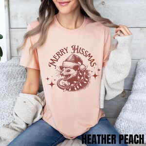 K&ouml;nnte beinhalten: Ein pfirsichfarbenes T-Shirt mit einem Aufdruck eines Opossums, das einen Weihnachtsmann-Hut und Weihnachtslichter tr&auml;gt. Der Text "Merry Hissmas" ist in einem Kreis um das Opossum geschrieben.