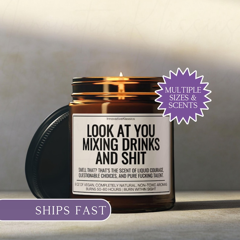 May include: A brown glass jar candle with a black lid, featuring the text "LOOK AT YOU MIXING DRINKS AND SHIT." The label also includes the scent description and burn time. A purple sticker says "MULTIPLE SIZES & SCENTS."