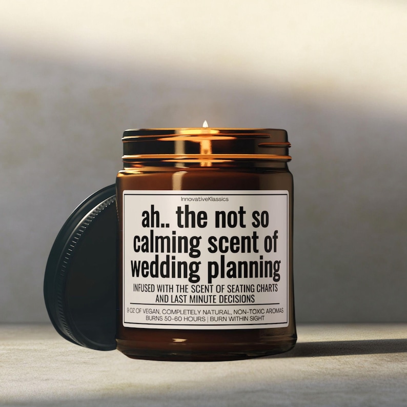 May include: A brown glass candle jar with a black lid. The candle is labeled "ah... the not so calming scent of wedding planning". The label also says "Infused with the scent of seating charts and last minute decisions".