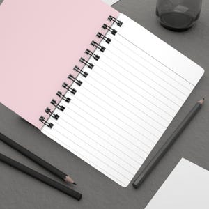 Op de afbeelding: Een roze en wit spiraalgebonden notitieboek met gelinieerde pagina's. Twee zwarte potloden liggen op een grijs oppervlak naast het notitieboek.