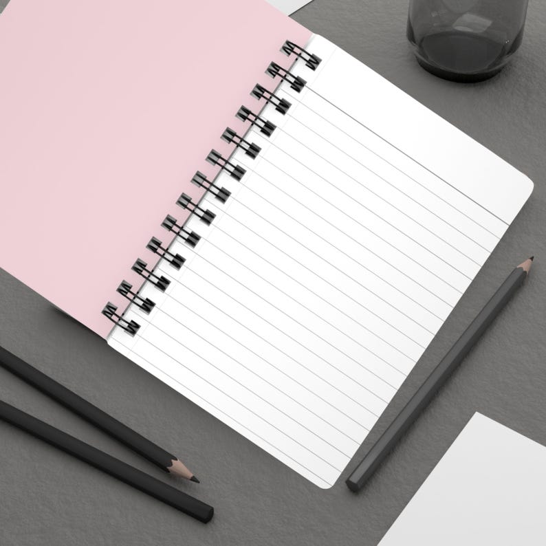 Op de afbeelding: Een roze en wit spiraalgebonden notitieboek met gelinieerde pagina's. Twee zwarte potloden liggen op een grijs oppervlak naast het notitieboek.