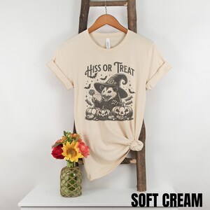 May include: A soft cream colored t-shirt with a graphic of a possum wearing a witch hat and holding a lollipop. The text "Hiss or Treat" is above the possum.