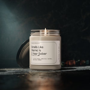 May include: A beige candle with a clear glass jar and a black lid. The candle has a white label with the text "Smells Like (Name) Is 1 Year Sober" and a small heart symbol. The text "So Proud of You" is printed below the main text. The label also includes the text "9 oz of Vegan, Completely Natural, Non-Toxic Aromas".