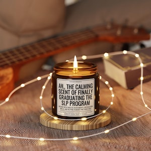 Op de afbeelding: Een brandende kaars in een bruine glazen pot met een gouden deksel, op een houten onderzetter. Het etiket luidt "AH.. THE CALMING SCENT OF FINALLY GRADUATING THE SLP PROGRAM". Slingers en een ukelele staan op de achtergrond.