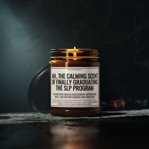 Op de afbeelding: Een brandende kaars in een bruine glazen pot met een zwart deksel. Op het etiket staat "AH.. THE CALMING SCENT OF FINALLY GRADUATING THE SLP PROGRAM." De kaars is veganistisch, natuurlijk en niet-toxisch, met een brandtijd van 50-60 uur.