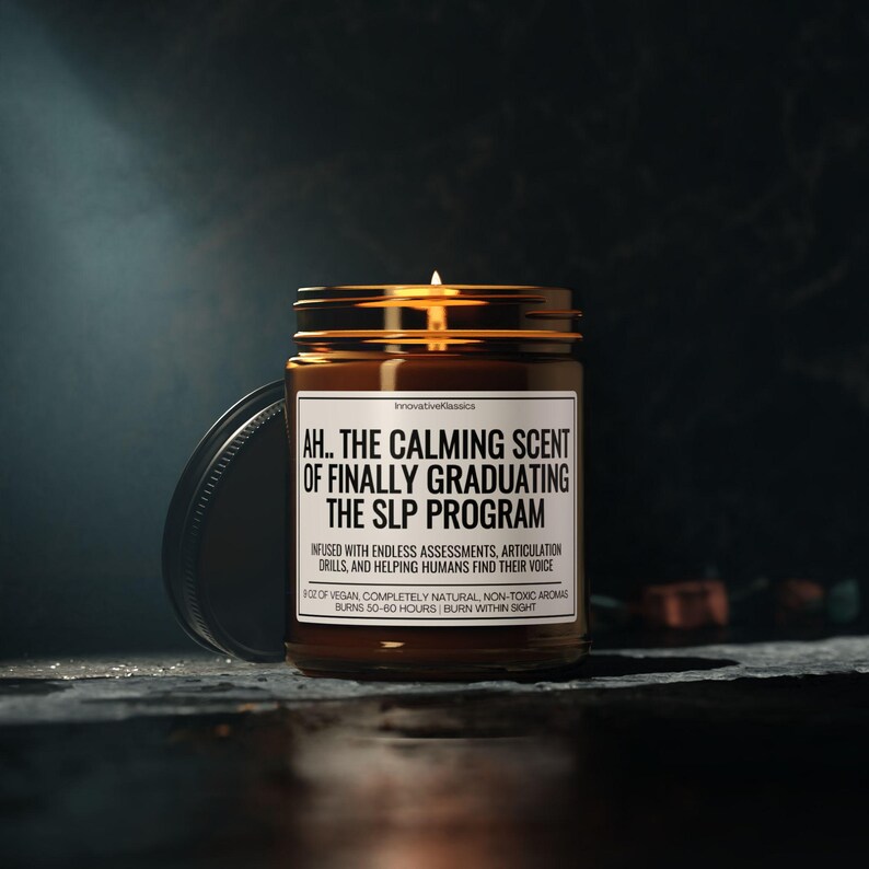 Op de afbeelding: Een brandende kaars in een bruine glazen pot met een zwart deksel. Op het etiket staat "AH.. THE CALMING SCENT OF FINALLY GRADUATING THE SLP PROGRAM." De kaars is veganistisch, natuurlijk en niet-toxisch, met een brandtijd van 50-60 uur.
