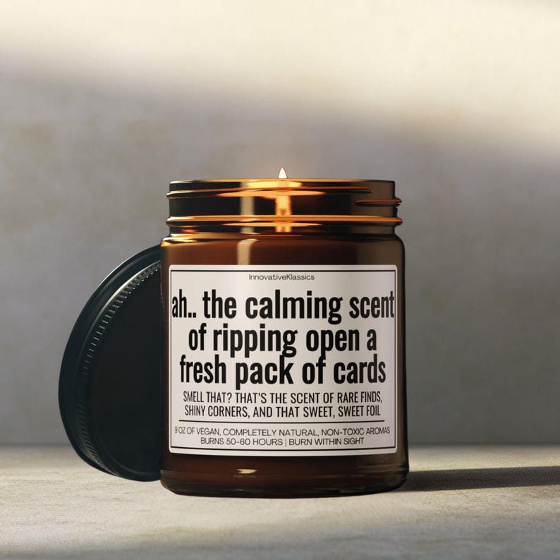 May include: A lit brown glass jar candle with a black lid. The label reads "ah.. the calming scent of ripping open a fresh pack of cards." The text below describes the scent as rare finds, shiny corners, and sweet foil. The candle burns for 50-60 hours.