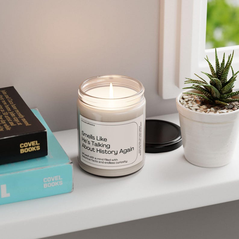 May include: A white candle with a clear glass jar and a black lid. The candle has a label that reads "Smells Like He's Talking About History Again" with the text "Infused with a mind filled with historical facts and endless curiosity".