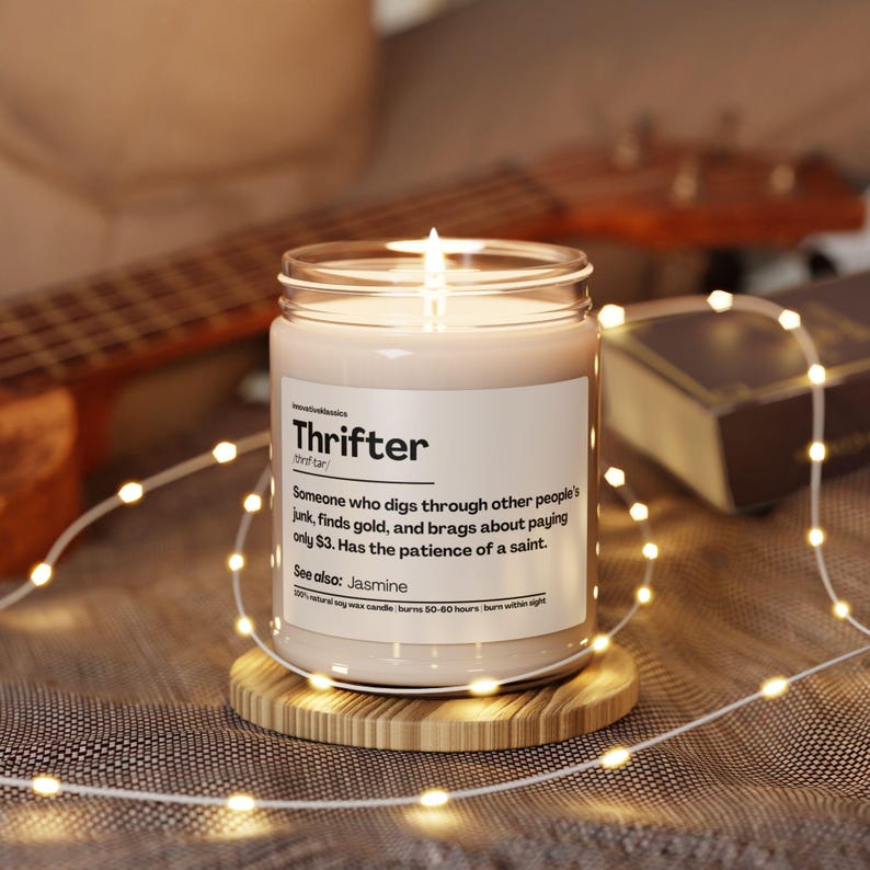 May include: A lit, white candle in a glass jar with the word "Thrifter" on the label. The label defines a thrifter. The candle sits on a wooden coaster, surrounded by string lights. The candle is a 100% natural soy wax candle.