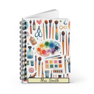 Puede incluir: Un cuaderno blanco de espiral con un dise&ntilde;o colorido de pinceles, paletas de pintura y otros materiales de arte. El cuaderno tiene una etiqueta amarilla con el texto "Sra. Smith" en &eacute;l.