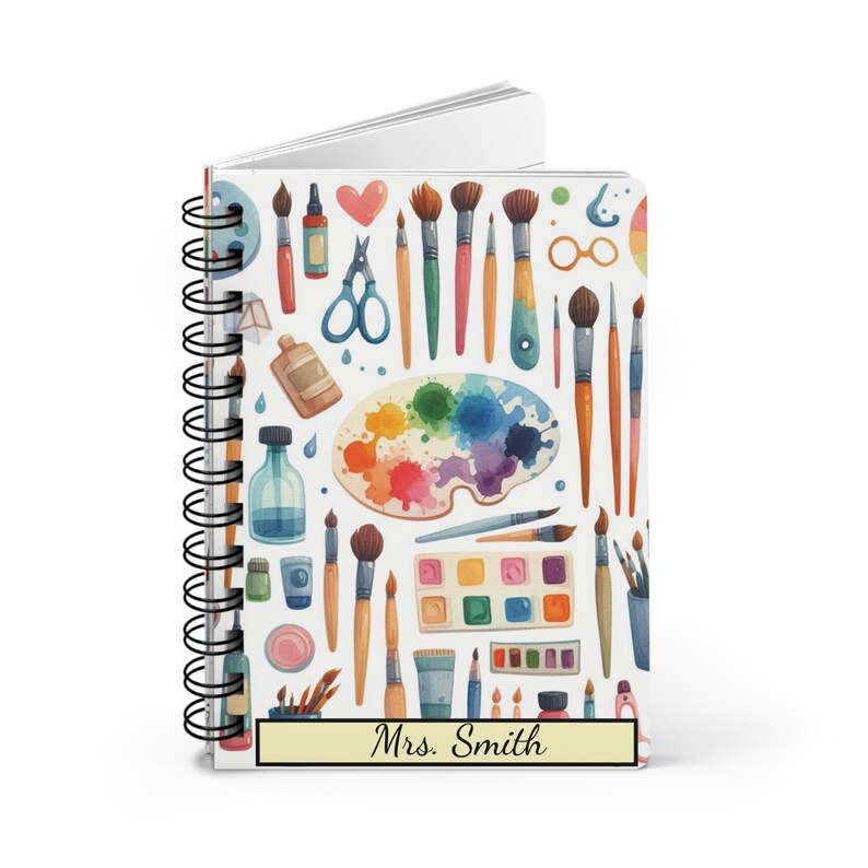 Puede incluir: Un cuaderno blanco de espiral con un dise&ntilde;o colorido de pinceles, paletas de pintura y otros materiales de arte. El cuaderno tiene una etiqueta amarilla con el texto "Sra. Smith" en &eacute;l.
