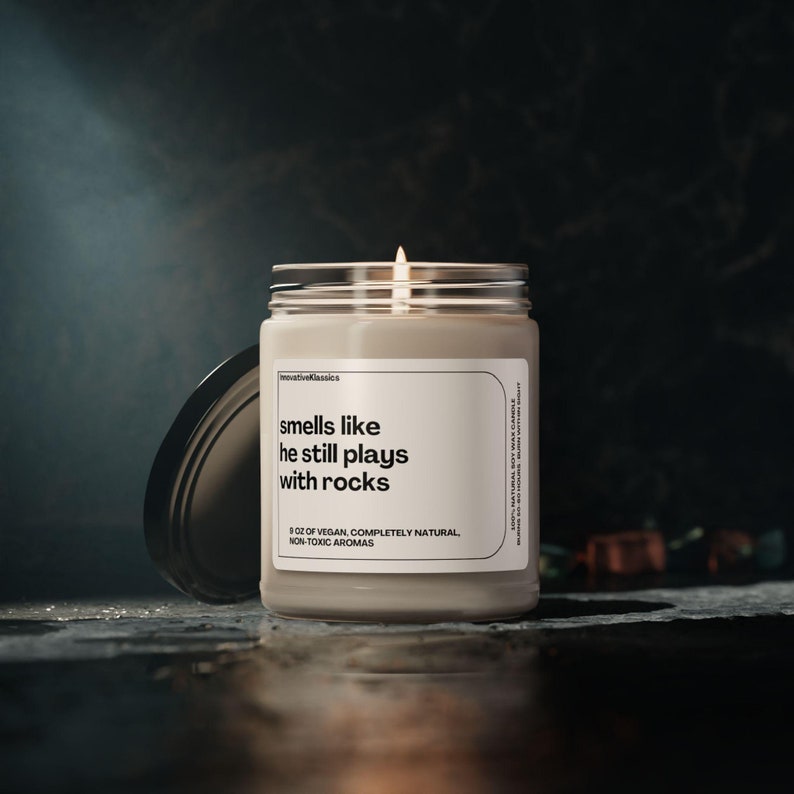 May include: A beige candle with a glass jar and a black lid. The candle has a white label with the text "smells like he still plays with rocks" and "8 oz of vegan, completely natural, non-toxic aromas".