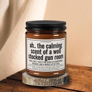 Puede incluir: Una vela en tarro de cristal marr&oacute;n con tapa negra, sobre una peque&ntilde;a rodaja de madera. La etiqueta dice "ah.. the calming scent of a well stocked gun room". El texto tambi&eacute;n menciona p&oacute;lvora, libertad y acero.