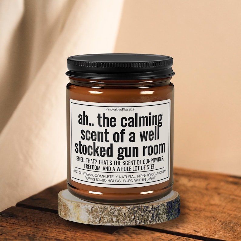 Puede incluir: Una vela en tarro de cristal marr&oacute;n con tapa negra, sobre una peque&ntilde;a rodaja de madera. La etiqueta dice "ah.. the calming scent of a well stocked gun room". El texto tambi&eacute;n menciona p&oacute;lvora, libertad y acero.