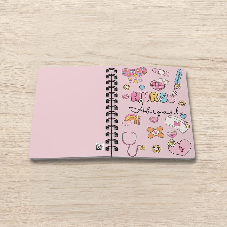 Op de afbeelding: Een roze spiraalnotitieblok met een witte omslag met een kleurrijk ontwerp van medische stickers en de tekst "NURSE Abigail".