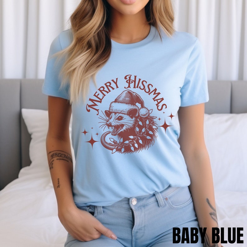 K&ouml;nnte beinhalten: Ein hellblaues T-Shirt mit einer roten Grafik eines Opossums, das einen Weihnachtsmann-Hut tr&auml;gt und von Weihnachtslichtern umgeben ist. Der Text "Merry Hissmas" ist in roter Kursivschrift um das Opossum herum geschrieben.
