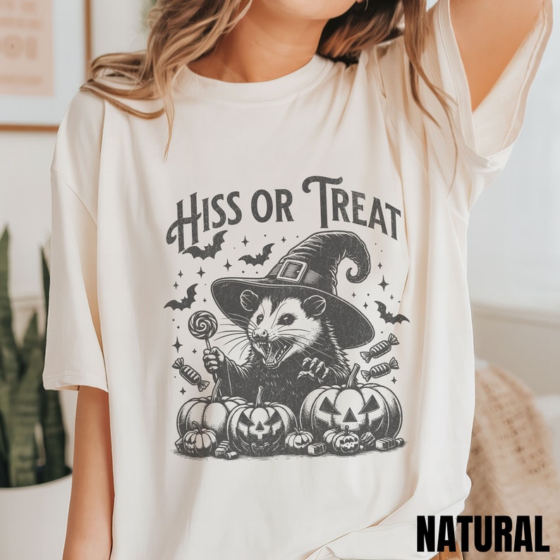 May include: A cream colored t-shirt with a black and white graphic of a possum wearing a witch hat and holding a lollipop. The text "Hiss or Treat" is above the possum. The t-shirt is labeled "NATURAL".