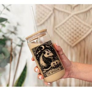 May include: A glass tumbler with a brown lid and a straw. The tumbler has a black and white illustration of an opossum with a pumpkin and the text "19 - THE OPOSSUM - 51".