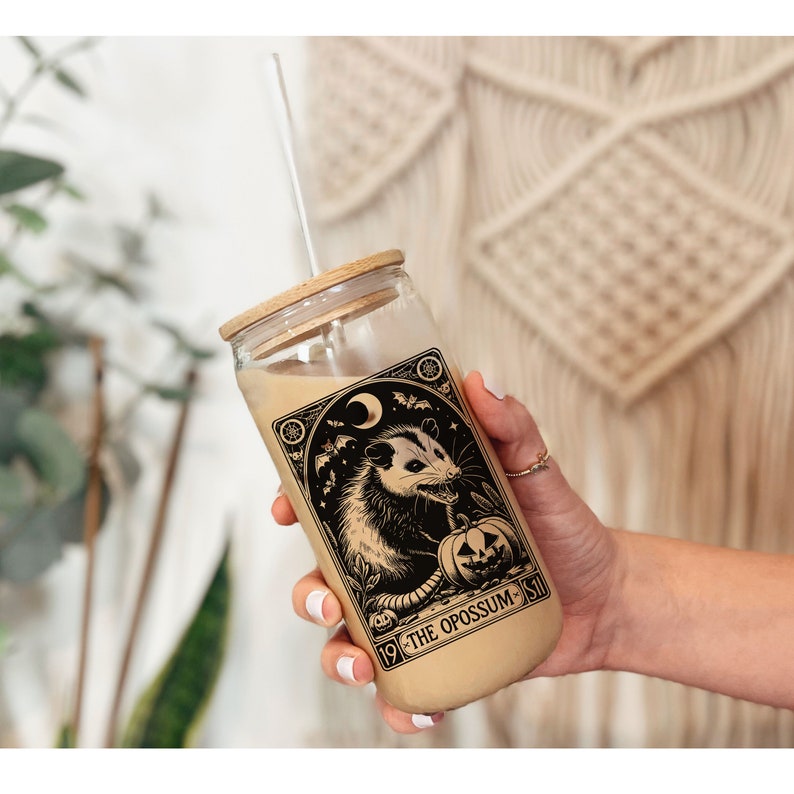 May include: A glass tumbler with a brown lid and a straw. The tumbler has a black and white illustration of an opossum with a pumpkin and the text "19 - THE OPOSSUM - 51".