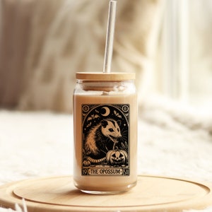 May include: A glass jar with a wooden lid and a straw. The jar has a black and white illustration of an opossum holding a jack-o-lantern. The text on the jar reads "19 - THE OPOSSUM - 11".