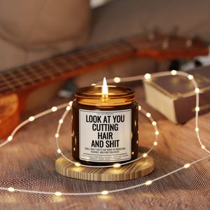 Puede incluir: Una vela encendida en un frasco marr&oacute;n con tapa dorada, sobre un posavasos de madera. La vela tiene una etiqueta blanca con el texto "LOOK AT YOU CUTTING HAIR AND SHIT". Un ukelele y luces de cadena est&aacute;n en el fondo.