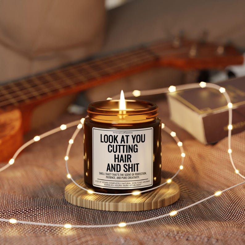 Puede incluir: Una vela encendida en un frasco marr&oacute;n con tapa dorada, sobre un posavasos de madera. La vela tiene una etiqueta blanca con el texto "LOOK AT YOU CUTTING HAIR AND SHIT". Un ukelele y luces de cadena est&aacute;n en el fondo.