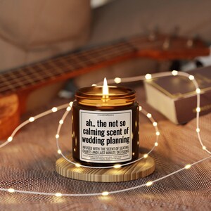 May include: A brown glass candle with a white label that reads "ah.. the not so calming scent of wedding planning". The candle is lit and sitting on a wooden coaster. The candle is surrounded by string lights.