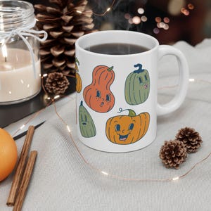 Op de afbeelding: Een witte keramische mok met een kleurrijk cartoonontwerp van pompoenen en pompoenen met gezichten. De mok heeft een zwart handvat en is gevuld met koffie.