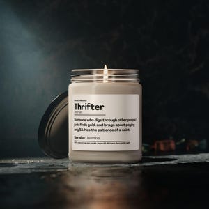 May include: A lit, white candle in a glass jar with a silver lid. The label reads "Thrifter" with a definition and "See also: Jasmine." The candle is on a dark surface.